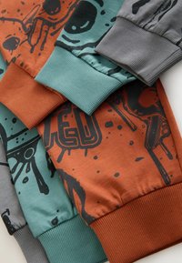 Drie langemouwen shirts in oranje, teal en grijs stoffen met gedurfde zwarte grafische prints en geribbelde manchetten in bijpassende kleuren.