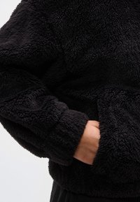 Άτομο που φοράει μαύρο, χνουδωτό μπουφάν fleece με το ένα χέρι μερικώς μέσα στην πλαϊνή τσέπη του μπουφάν.