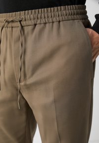 Rjave hlače z elastiko v pasu, z vrvico in stranskimi žepi, izdelane iz gladkega materiala s prilagojenim krojem, ki poudarja subtilno teksturo.