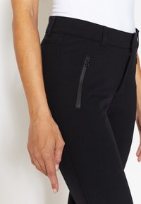 Schwarze stretchige Hose mit slim fit, ausgestattet mit einer Reißverschlusstasche an der Seite und einer glatten Textur. Der Bund ist für einen sicheren Sitz konzipiert.