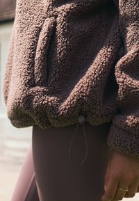 Pullover in pile marrone con una superficie strutturata e pelosa e un elastico con cordino all'orlo; abbinato a collant lisci e aderenti.