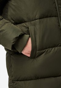 Casaco acolchoado verde oliva com uma textura suave, apresenta um punho canelado e um bolso lateral, exibindo um design acolchoado para isolamento.