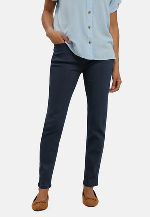 Kvinde iført marineblå smalle bukser, lyseblå skjorte med knapper og brune flade sko, stående med den ene hånd i lommen.