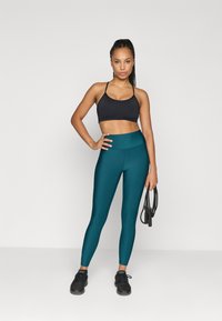 Μαύρο αθλητικό σουτιέν, ψηλής μέσης τιρκουάζ κολάν και μαύρα αθλητικά παπούτσια. Το μοντέλο κρατάει ένα σχοινί σκαρφαλώνοντας, και στέκεται μπροστά σε λευκό φόντο.