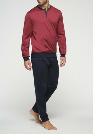 Uomo che indossa una maglietta henley rossa a maniche lunghe con bordino nero e pantaloni da jogging neri, in piedi a piedi nudi su uno sfondo semplice.