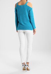 Pull en tricot bleu à décolleté asymétrique, associé à un jean blanc ajusté et des chaussures à talons argentées. Texture lisse, style décontracté.