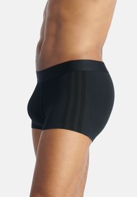 Torso masculino y parte superior de las piernas con calzoncillos ajustados negros de tipo bóxer con tres franjas verticales oscuras en el lateral, sobre un fondo blanco.