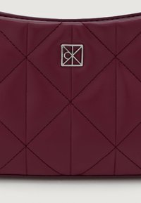 Μαρόν τσάντα από καπιτονέ ψεύτικο δέρμα με διαγώνια ραφή, ασημένιο λογότυπο και λείο υλικό. Συμπαγές ορθογώνιο σχήμα.