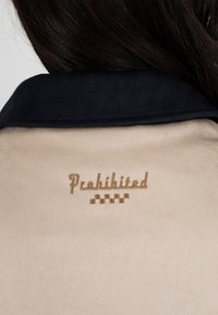 Personne aux cheveux foncés portant une veste beige avec un col noir et le mot brodé en or "Interdit" ainsi qu'un motif à carreaux dans le dos.