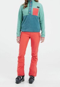 Polarowy sweter w odcieniach niebieskiego i zielonego z koralową kieszenią na piersi, zestawiony z dopasowanymi jasnopomarańczowymi spodniami i czarnymi butami narciarskimi.