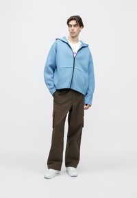 Ανοιχτό μπλε φούτερ με φερμουάρ και μεγάλη τσέπη μπροστά, συνδυασμένο με καφέ παντελόνι καρό και λευκά αθλητικά παπούτσια. Ομαλή υφή και άνετη γραμμή.