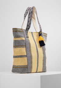 Borsa a tracolla a righe in tessuto giallo, nero e bianco. Caratterizzata da due manici robusti e un tassello decorativo. Superficie testurizzata con motivi distintivi.