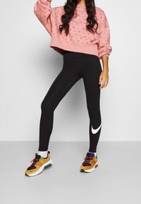 Rosa sweatshirt med präglat cirkelmönster, svarta leggings med en vit Nike-swoosh, och färgglada sneakers. Avslappnad atletisk outfit.