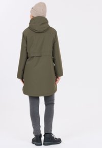 Olivgrön vattentät jacka med huood, medellång design och en flapdetalj på baksidan. Bärs med grå jeans och svarta stövlar.