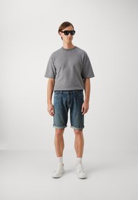 Tricou gri cu mânecă scurtă, pantaloni scurți din denim albastru cu margini răsucite, adidași albi și ochelari de soare negri pe un fundal simplu.