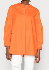 Haut long orange à col montant, avec un devant plissé et des manches froncées. Porté sur un jean blanc déchiré. Texturé lisse.