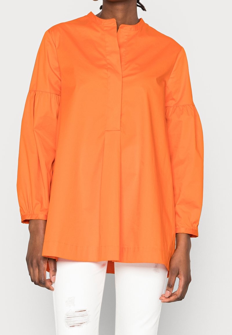 Haut long orange à col montant, avec un devant plissé et des manches froncées. Porté sur un jean blanc déchiré. Texturé lisse.