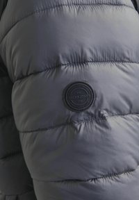Chaqueta de plumas gris con textura acolchada, que presenta un parche circular negro con la etiqueta "JACK & JONES" en el lado izquierdo.