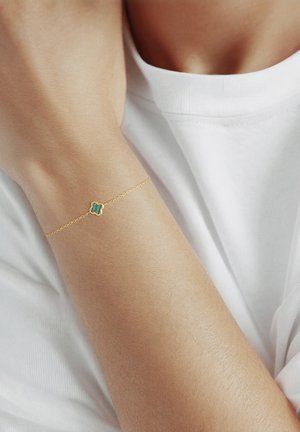 Bracelet délicat en chaîne d'or avec un petit porte-bonheur en forme de trèfle vert porté au poignet d'une femme par-dessus une chemise blanche.