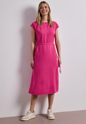 Uśmiechnięta kobieta stojąca przy ścianie, ubrana w jasnoróżową sukienkę midi z troczkami po bokach i beżowo-białe trampki.