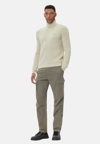 Maglione a collo alto in maglia beige chiaro abbinato a pantaloni cargo oliva. I pantaloni hanno molte tasche. Scarpe nere con lacci completano l'outfit.
