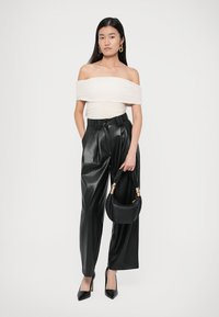 Off-shoulder crèmekleurig top met geplooide textuur, hoge taille zwarte nepleren wijde broek, puntige zwarte hakken en een kleine zwarte handtas.