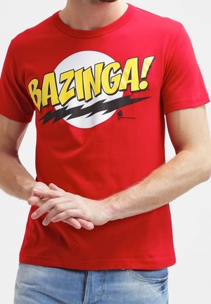 Rotes Kurzarmshirt mit dem Schriftzug „BAZINGA!“ in gelben Buchstaben über einem weißen Kreis und schwarzen Blitzsymbolen, getragen von einer Person mit verschränkten Händen.