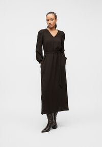 Γυναίκα που φοράει μακρύ μαύρο φόρεμα με ζώνη και μακριά μανίκια, καθώς και μαύρα μυτερά μποτάκια με ψηλό τακούνι, στέκεται μπροστά σε απλό φόντο.