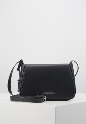 Czarna skórzana torba na ramię Calvin Klein z regulowanym paskiem i srebrnym logo z przodu, wyeksponowana na białym postumencie.