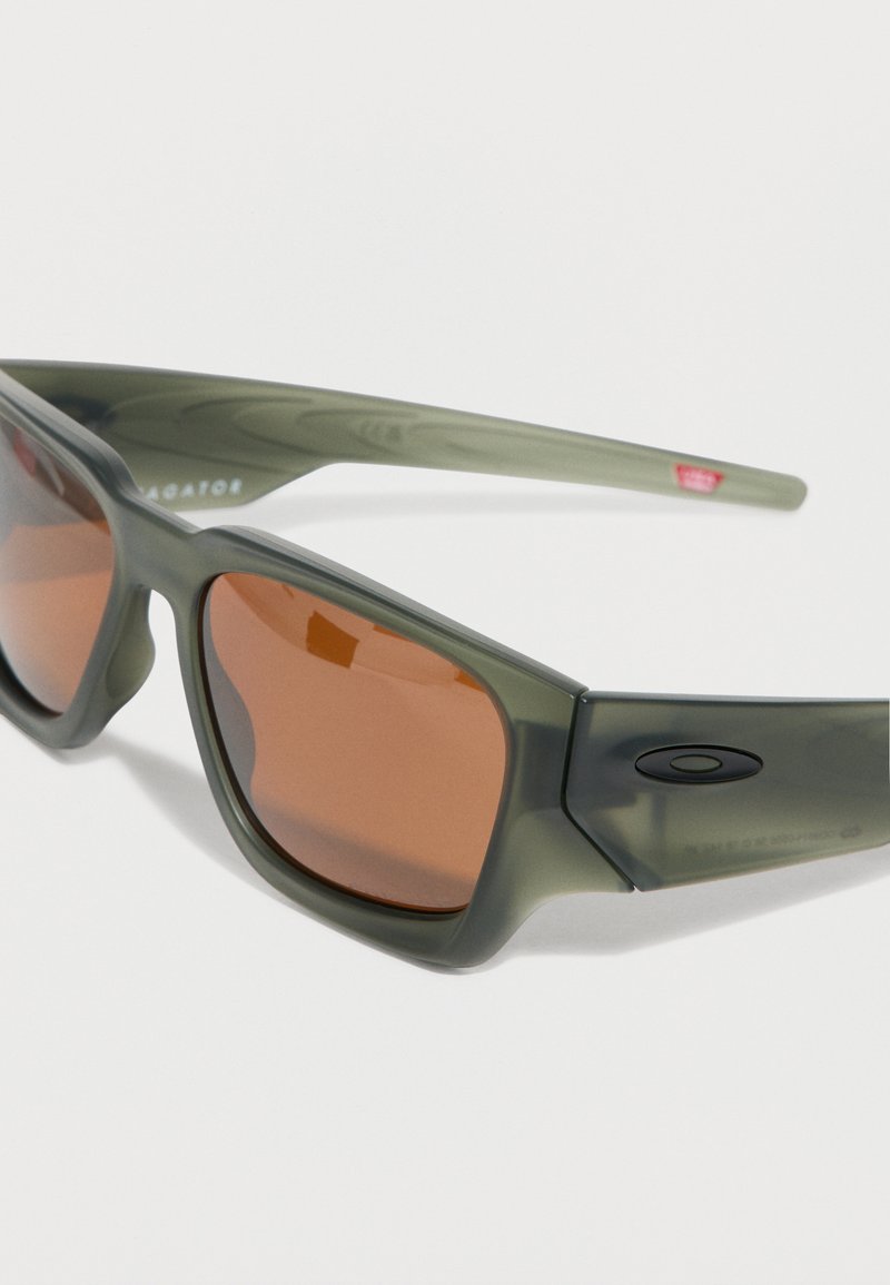 Quadratische Sonnenbrille mit grünen, durchsichtigen Rahmen und braun getönten Gläsern, mit einem kleinen schwarzen Logo am Bügel.