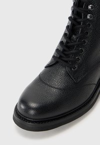 Bota de cuero negro hasta el tobillo con acabado texturizado, puntera redondeada, cordones negros y ojales metálicos. Suela resistente con detalle de costura en contraste.