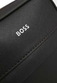 Svart texturerad läderväska med en silver "BOSS"-logotyp, mörkröd sömnad och en elegant design med distinkta vinklar.