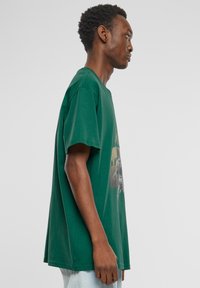 Grøn bomulds t-shirt med korte ærmer, med et grafisk tryk på forsiden. Løs pasform, rund hals og lige kant.