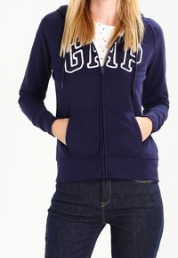 Sudadera con cremallera de color marino con letras blancas de "GAP", bolsillos tipo canguro y corte estructurado. Fabricada en un tejido suave y texturizado. Combinada con unos jeans oscuros.
