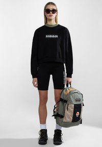 Sudadera negra con logo, pantalones cortos de ciclista negros, mochila gris y beige con bolsillo de malla, y botas de senderismo negras. Las gafas de sol completan el look.