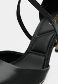 Nærbilde av en svart skinnhøyehælssandal med polstret innersåle og tynn ankelstropp på hvit bakgrunn.