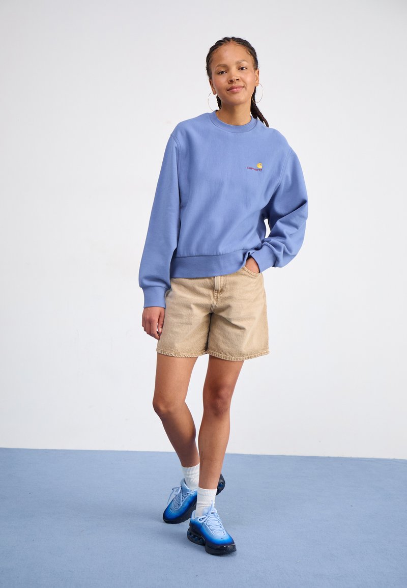 Sweatshirt bleu clair avec des poignets côtelés et un petit logo, associé à un short en denim beige. Le footwear se compose de baskets bleues et de chaussettes blanches.