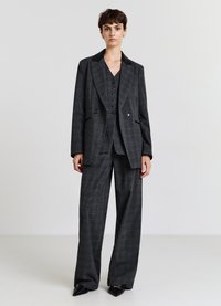 Donna che indossa un completo tre pezzi a scacchi grigio scuro con pantaloni a gamba larga e scarpe nere con la punta, in piedi contro uno sfondo semplice.