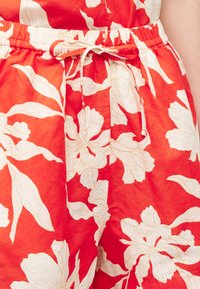 Κόκκινα υφασμάτινα σορτς με μεγάλο λευκό λουλουδάτο σχέδιο και ελαστική μέση δεμένη με λεπτό κόκκινο κορδόνι.