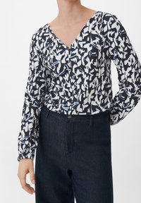 Persona che indossa una blusa a V con un motivo astratto bianco, blu e nero, con maniche lunghe e voluminose, infilata in jeans scuri a vita alta.