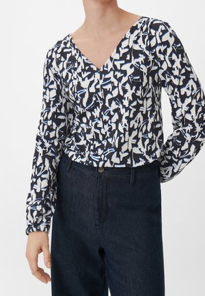Personne portant un haut en V à motifs abstraits noir, blanc et bleu, avec des manches longues bouffantes, rentré dans un jean sombre taille haute.