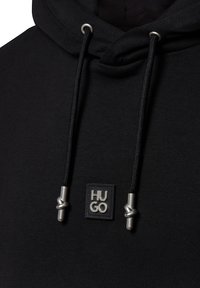 Felpa nera con cappuccio e cordino, dotata di accessori metallici. Include un'etichetta quadrata con il logo "HUGO" in grigio testurizzato. Tessuto morbido.