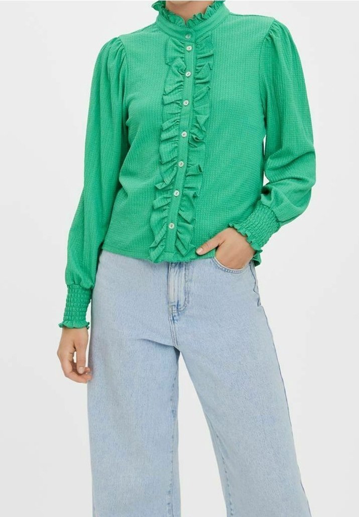 Femme portant un chemisier vert vif à manches longues avec des volants sur le devant et des poignets smockés, associé à un jean taille haute bleu clair.