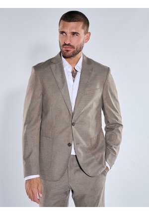 Mann mit kurzen Haaren und Bart, trägt einen hellbraunen Anzug und weißes Hemd, steht mit einer Hand in der Tasche und blickt zur Seite.