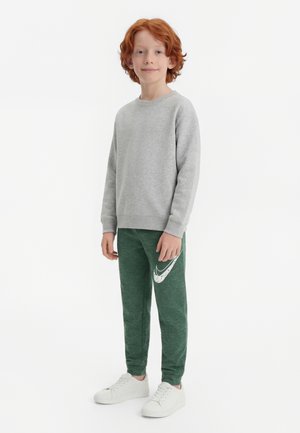 Băiat roșcat în picioare, purtând un hanorac gri, pantaloni de trening Nike verzi cu logo alb și adidași albi pe fundal alb.