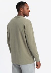 Uomo che indossa una maglietta a maniche lunghe verde oliva e pantaloni grigio chiaro, rivolto di lato su uno sfondo bianco semplice.