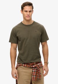 Oliven grøn bomulds t-shirt med rund hals, korte ærmer, der har et lille logo. Brunternet skjorte bundet om livet og lyse khaki bukser.