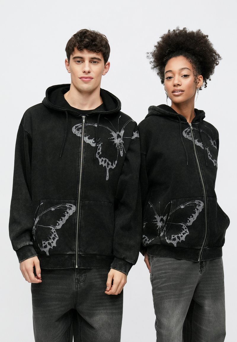 Melni rāvējslēdzēja sporta džemperi no mīksta auduma, ar lieliem pelēkiem taureņu attēliem priekšpusē un kabatās. Abi modeļi valkā pelēkas džinsa bikses.