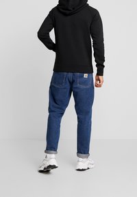 Sudadera negra con capucha y pantalones de mezclilla azules con los puños enrollados. Las zapatillas son blancas con acentos negros. Vista trasera sobre un fondo neutro.
