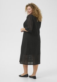 Robe noire midi avec un motif en chevrons, présentant des manches 3/4 et une texture translucide. Associée à des ballerines noires. Vue de dos.